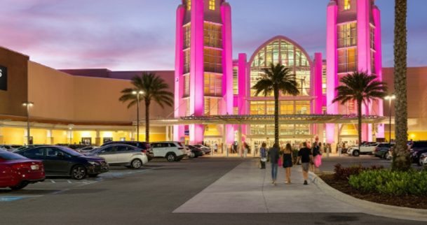 Sawgrass Mills mall entrance lit up with pink lights at dusk, featuring palm trees, parked cars, and shoppers heading inside for Black Friday.