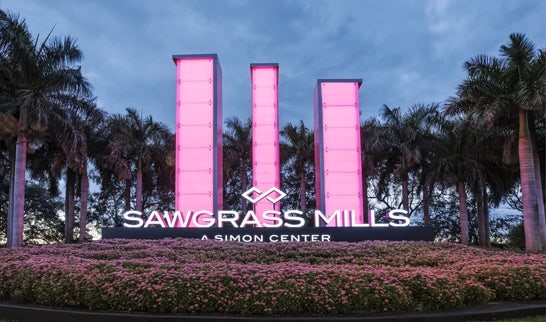 Main entrance sign of Sawgrass Mills mall illuminated in pink with palm trees in the background.